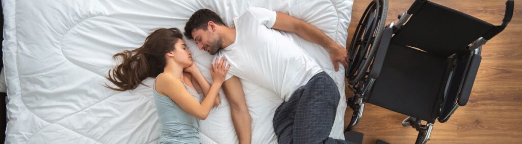 A young couple lays in bed facing each other. A wheelchair is sitting along the side of the bed indicating one of them has a disability. 