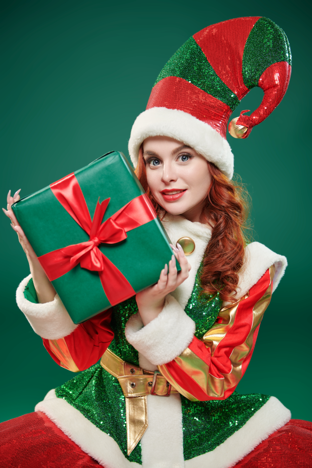 A young redheaded woman in a cute elf outfit, posing with a present against a green background. 