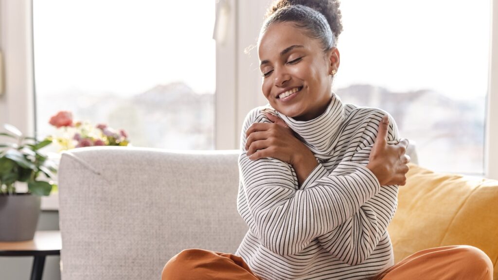 A woman of color sits on a couch with eyes closed and hugs herself