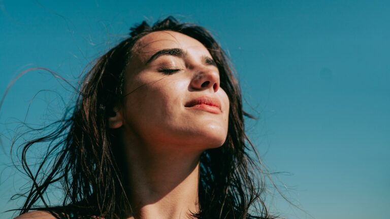 A young woman with her eyes closed and hair down looks up with a small smile in front of a blue background.