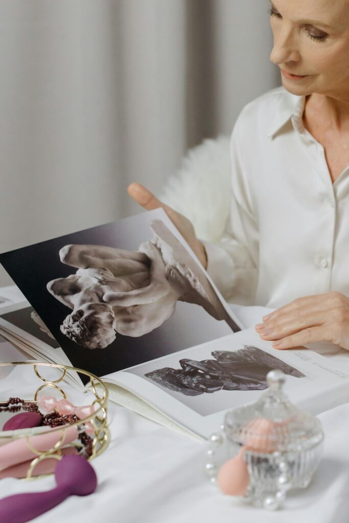 An older white woman, surrounded by sex toys, is looking through a book with nude greek statues. 