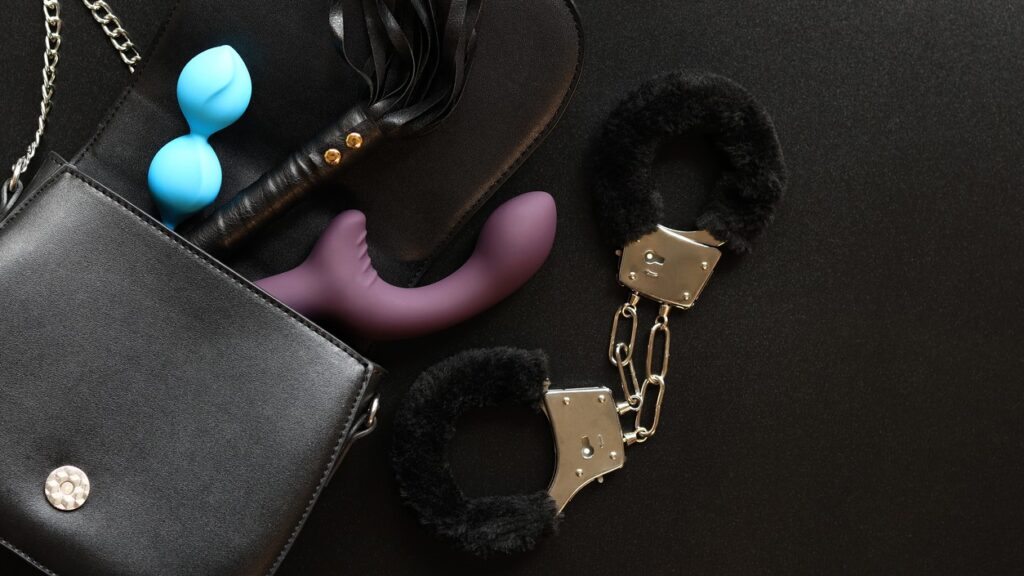 black furry handcuffs next to a black purse with sex toys and a black whip on a black background
