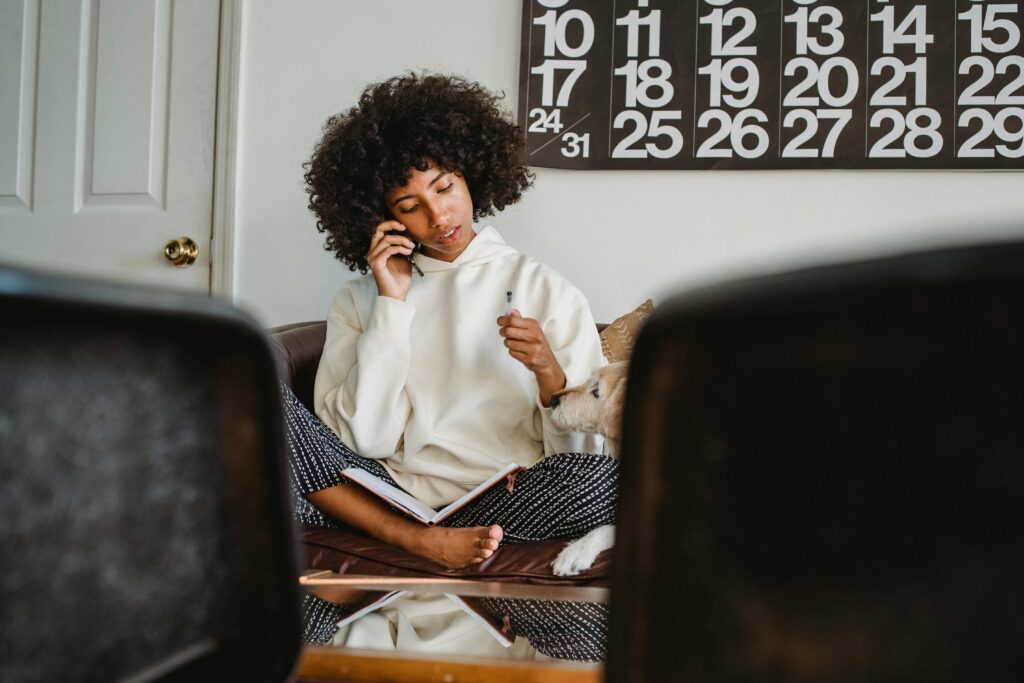 A woman with afro hair is sitting cross-legged on her couch. Talking on the phone. 