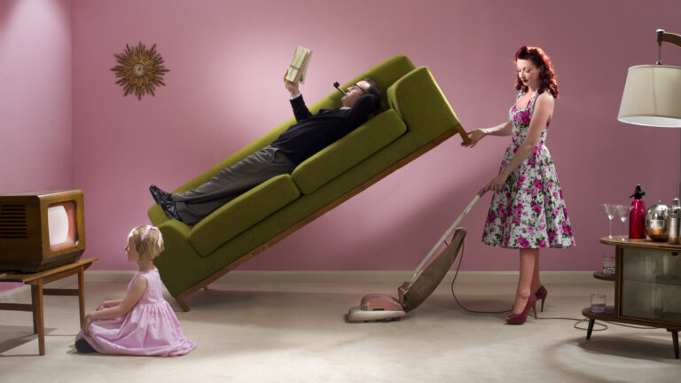 1950s style wife cleans around husband and child while husband lounges on a green couch.