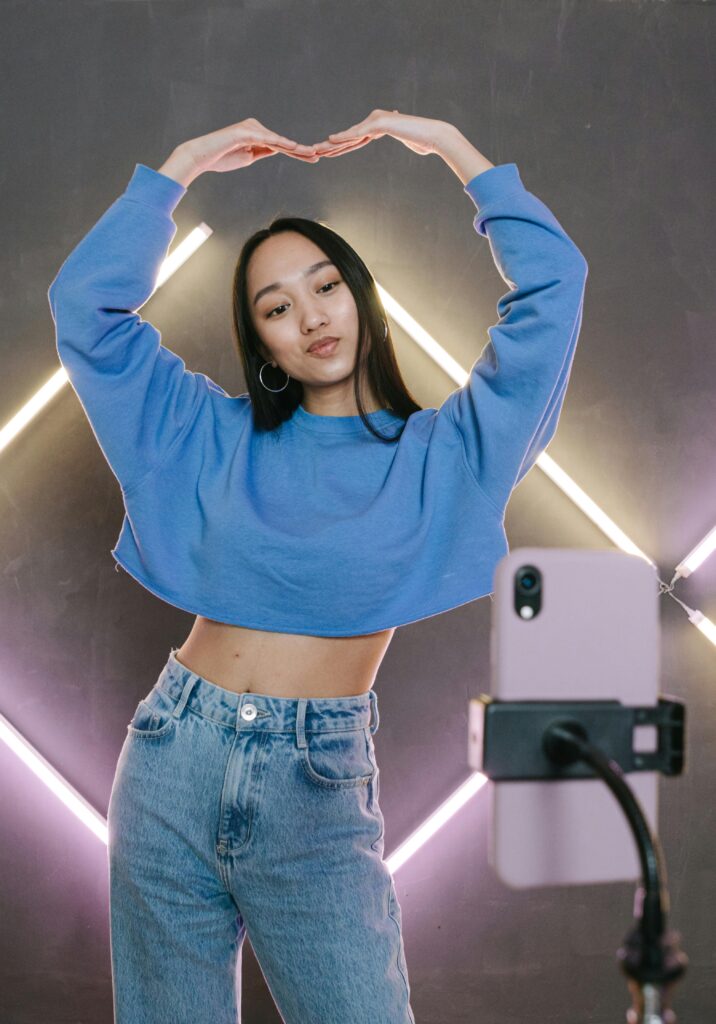 An Asian female standing in front of her phone, which is attached to a tripod phone stand. She has her arms in the air making a heart shape. 