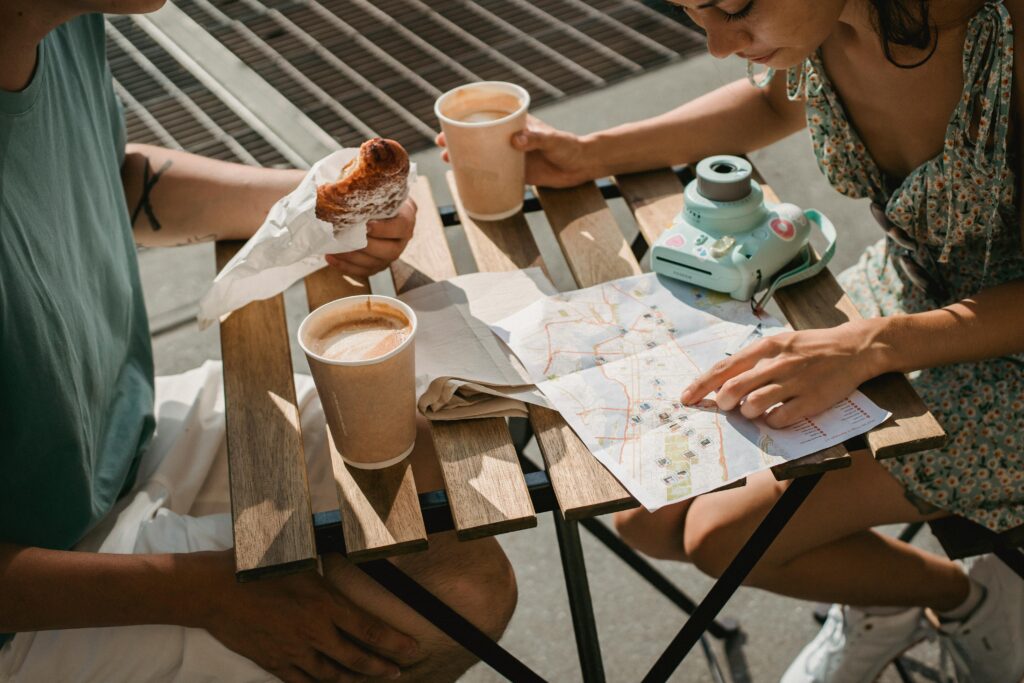 A couple eating outside at a cafe, looking at a roadmap. They have a camera. Traveling together allows couples to share memories. 