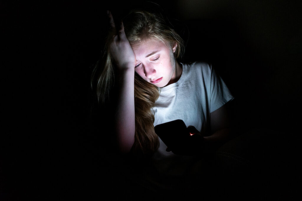 A young girl looks distraught looking at her phone possibly from image-based sexual abuse including cyberflashing or unwanted sexual content. 