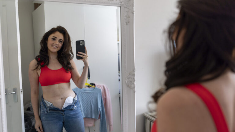 Over the shoulder shot of a woman with a stoma bag looking into a mirror in her bedroom in the North East of England. She is taking a selfie on her mobile phone, smiling at her reflection.