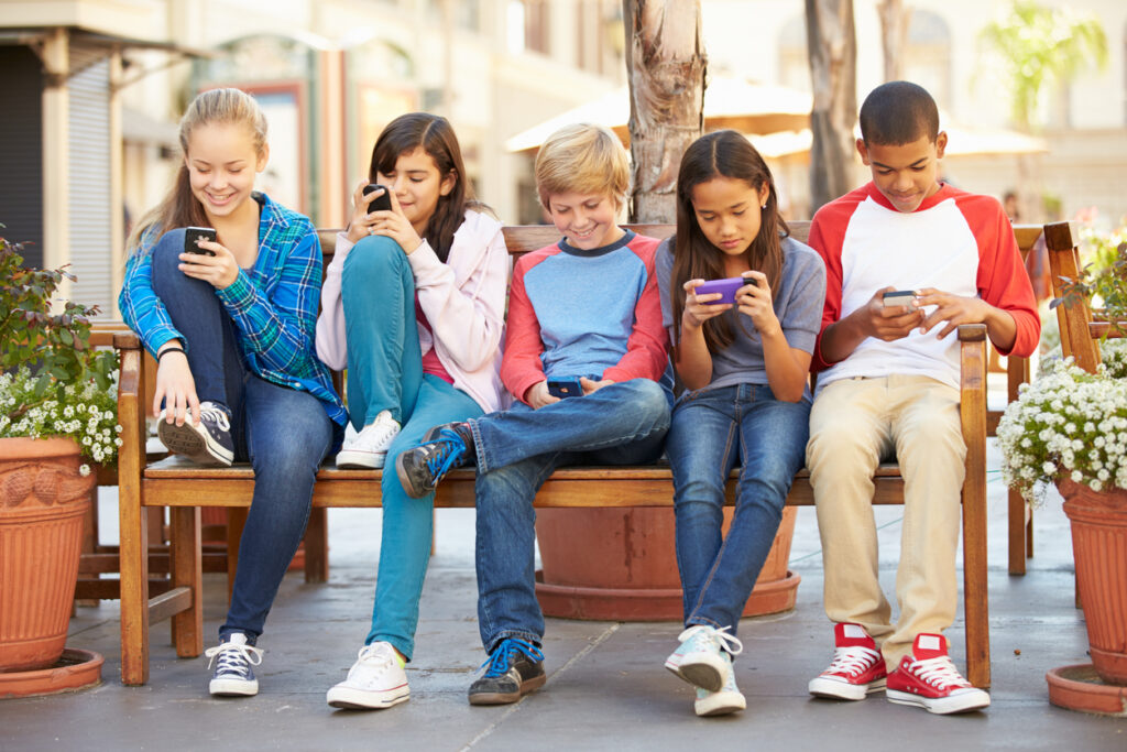 A group of tweens sitting on bench all looking at their cellphones. 