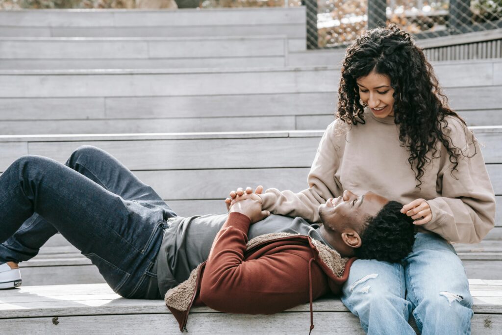 A young diverse couple are hanging out on a set of cement bleachers. The male is resting his head in the female's lap. They are looking at each other smiling. 