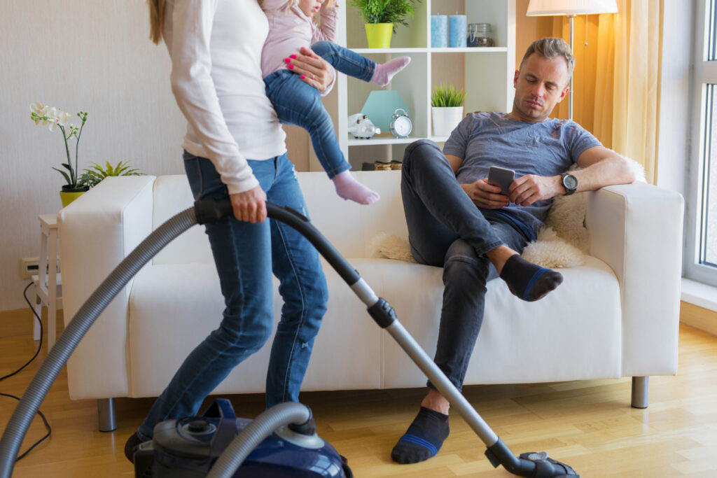 A woman holds her baby and vacuums a living space while her male partner sits on the couch scrolling through his phone. 