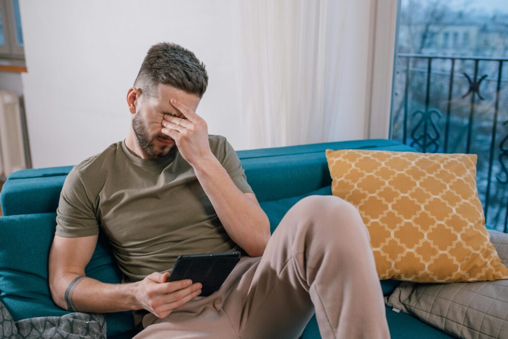 A man sits on a couch with his hand rubbing his eyes. He looks stressed. 