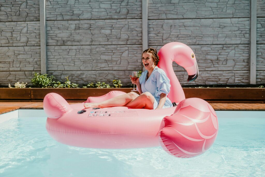 Young lady looking joyous in her pink flamingo pool toy floating in a pool with a glass of wine. 