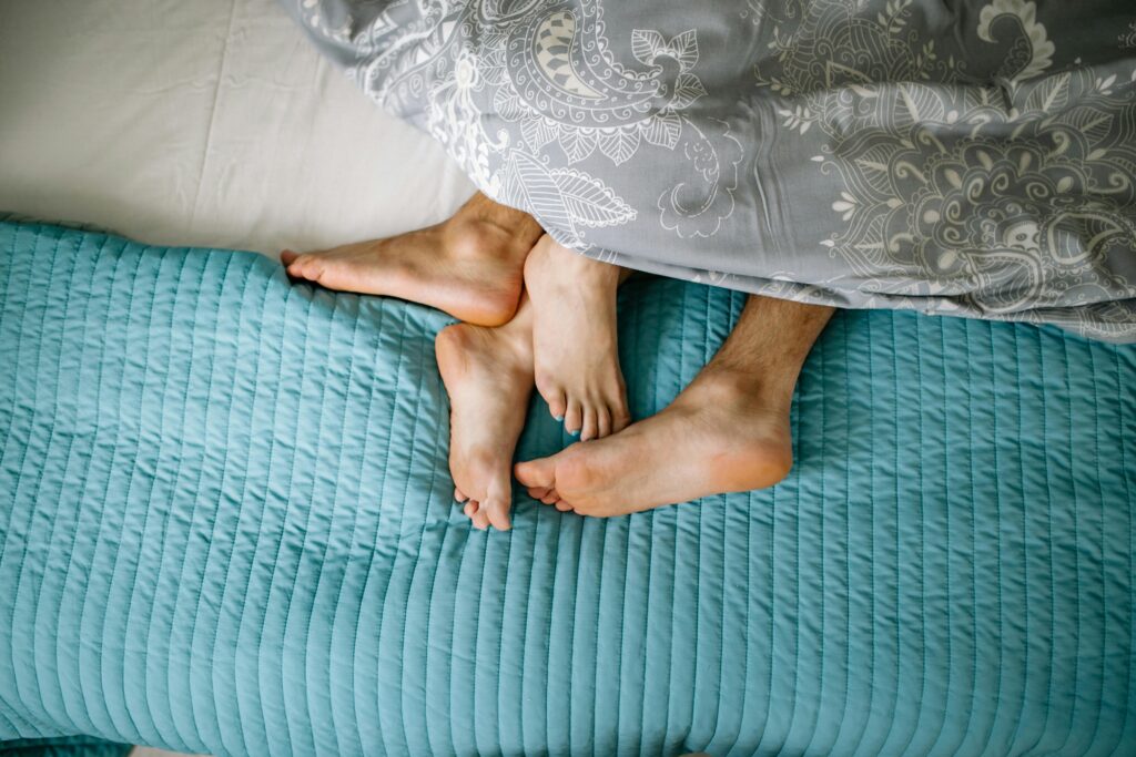 A pair of feet sticking out from under the covers of a bed. The bedding is light blue. 