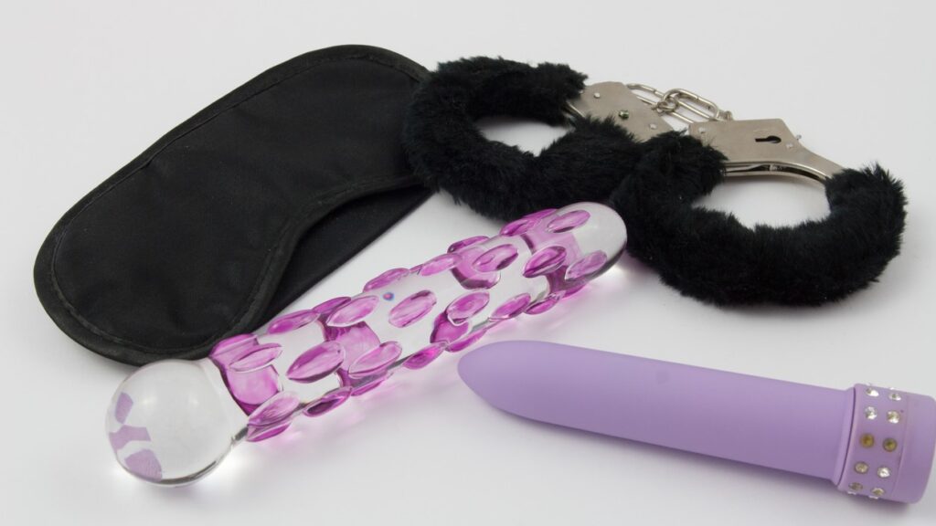 A black eye mask, black furry handcuffs, a purple and clear glass dildo and a purple vibrator with jewels at the base represent kinky sex toys