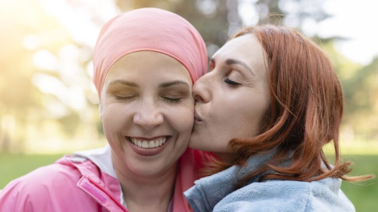 Lesbian couple, one is a cancer survivor with scarf on head being kissed on the cheek by her partner