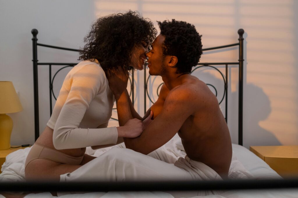 young african american couple in sitting up in bed, facing each other. They are both smiling with their faces close together like they are about to kiss. 