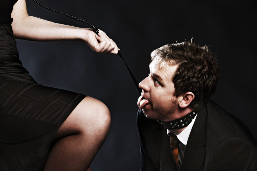 a female with a man in a suit on a leash with collar. the man is panting like a puppy dog.