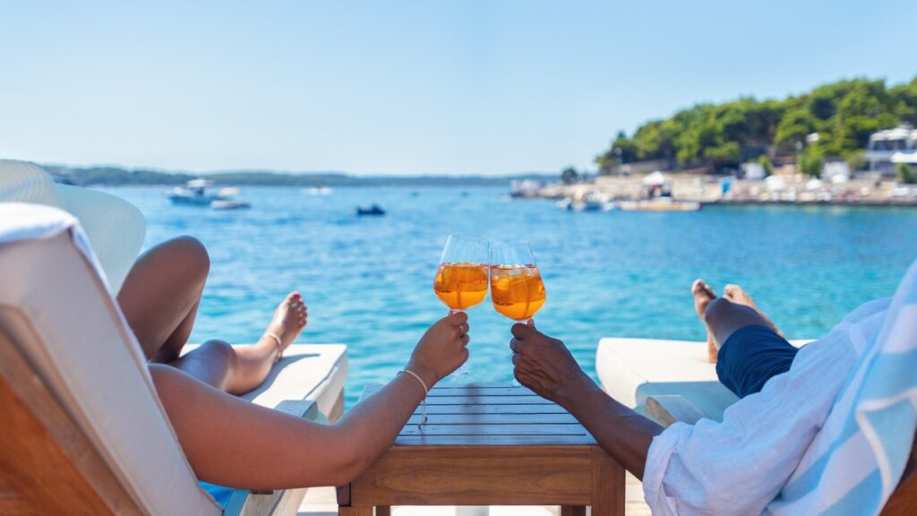 Couple on a tropical beach on lounge chairs seen from behind looking at the water while holding cocktails