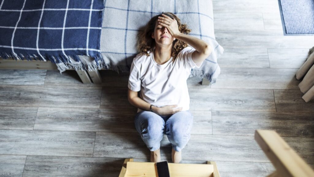 Young woman on the floor beside a bed holding her head and lower abdomen in pain from vulvodynia