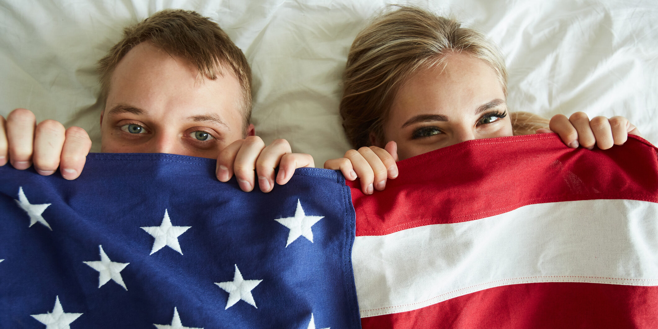 young couple peeking out under an american flag. It appears they are laying in a bed together.