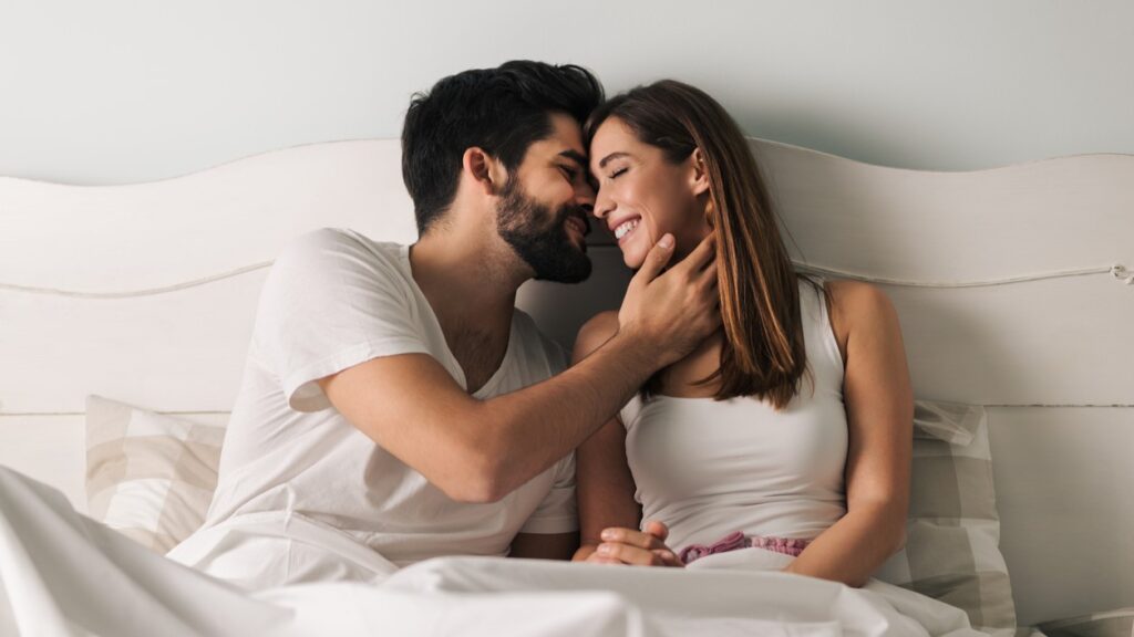 Young couple in bed smiling and leaning in for a kiss