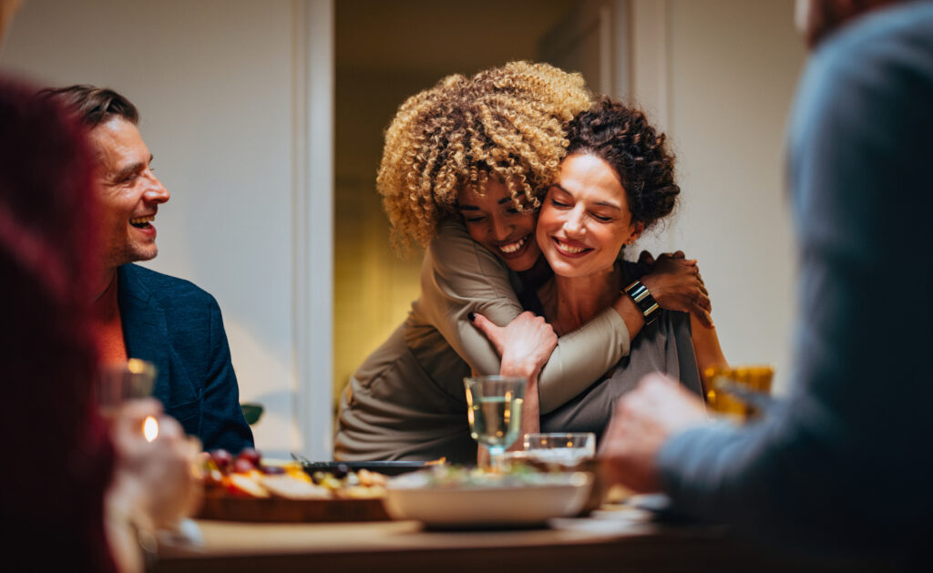 A woman hugs another woman from behind at a dinner table. 