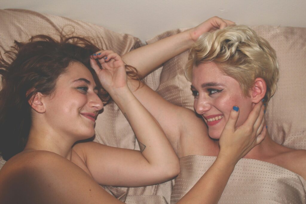 female female queer couple laying in bed together, smiling.