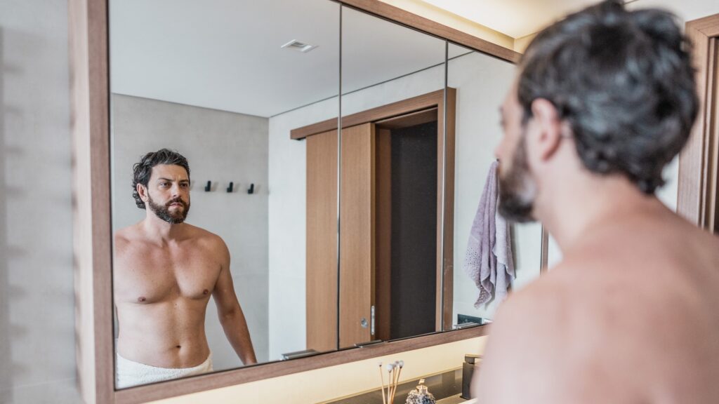 man in towel looks in mirror in bathroom