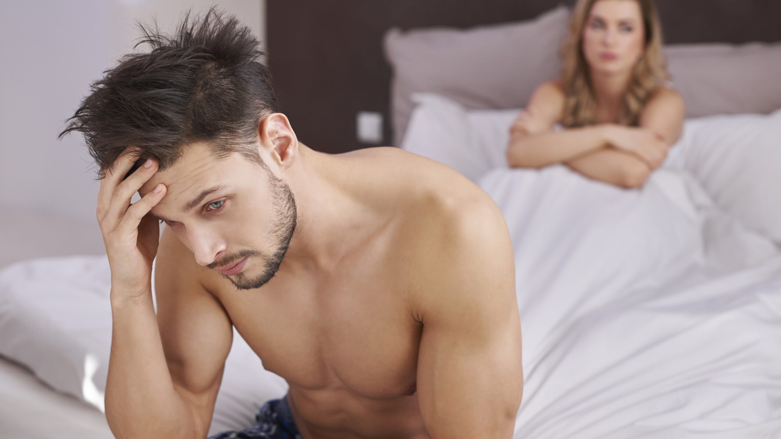 man sitting topless at edge of bed holding his head in frustration in front of female partner in bed looking disapointed