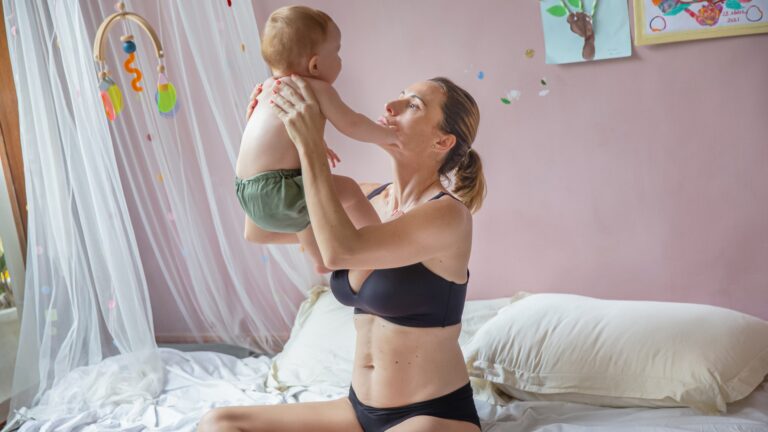 woman postpartum wears black bra and panties while holding up newborn baby
