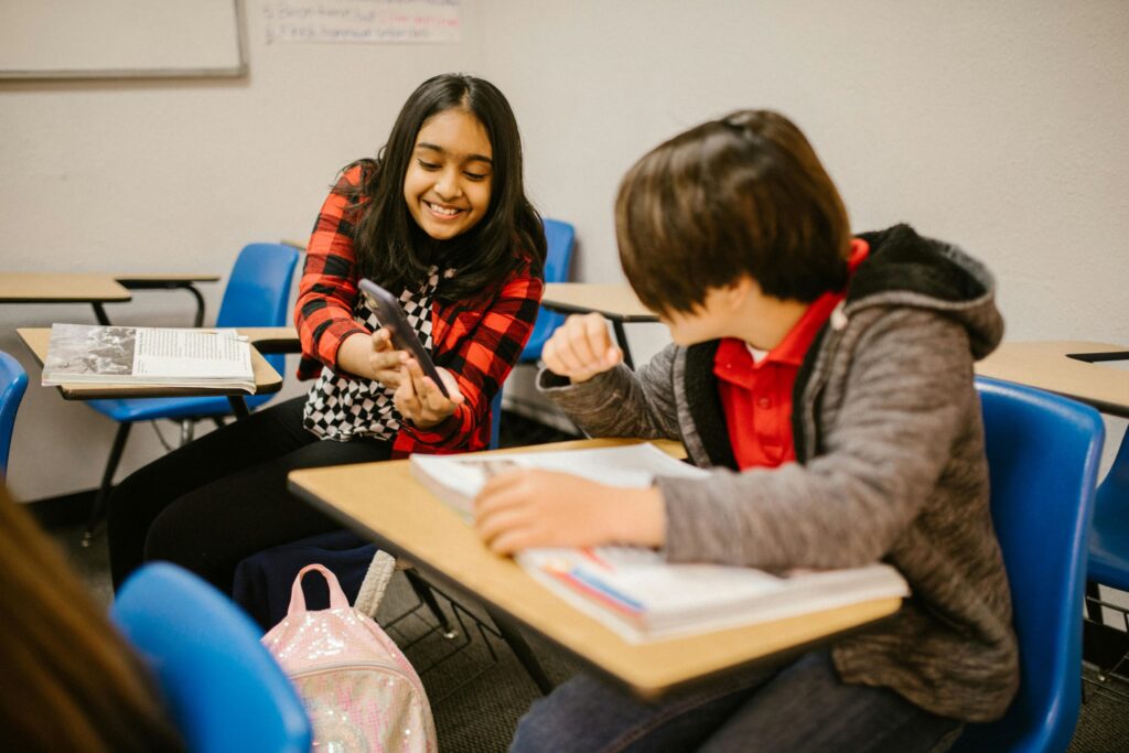 A young girl with a smartphone leans over in her desk and shows a young boy something on her phone inside a classroom.