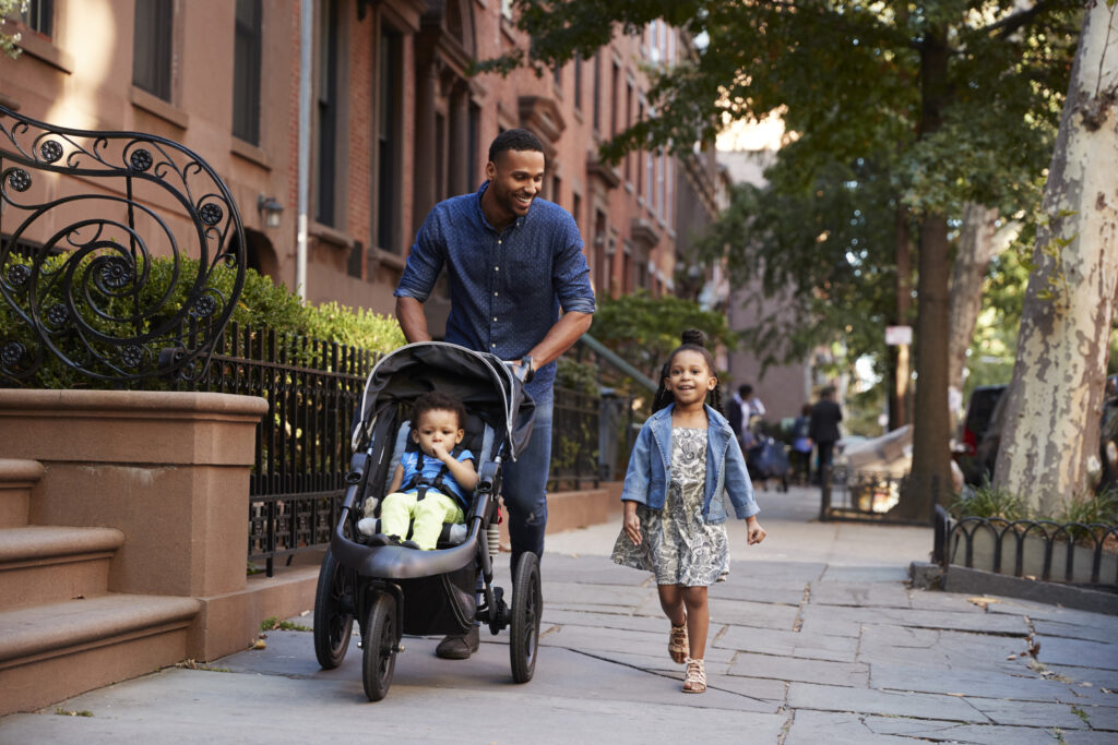 Diverse man walking a stroller with a baby in it down a city sidewalk, a toddler walks along the side.
