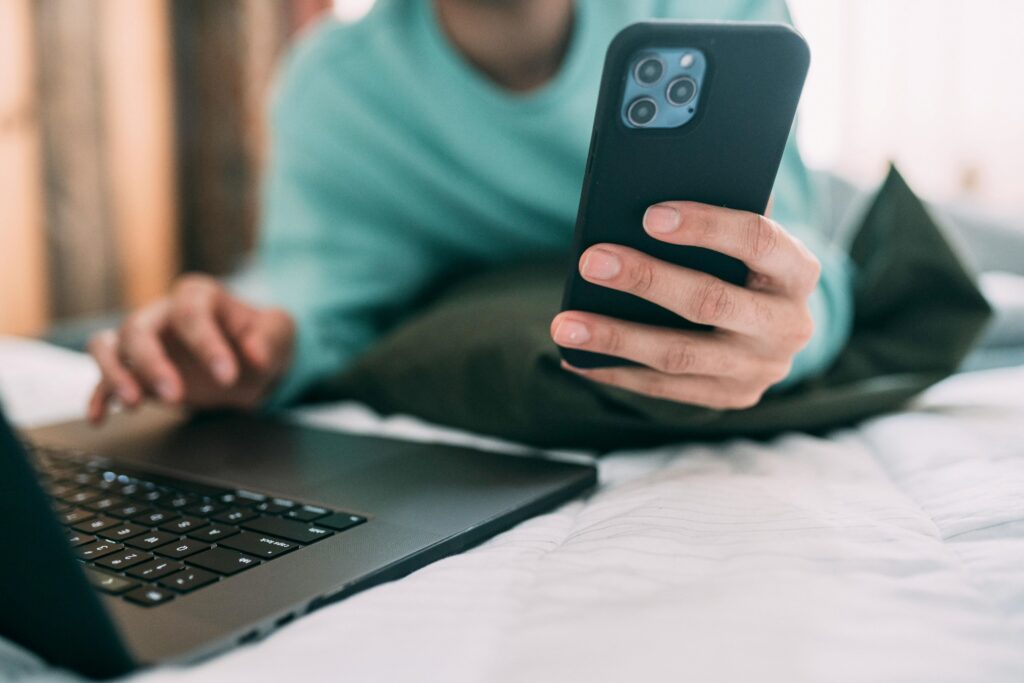 close up of a man's arms and hands using a laptop and cellphone at the same time while lying on a bed.