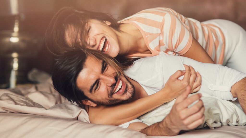 A happy young heterosexual couple smiling and cuddling in bed.